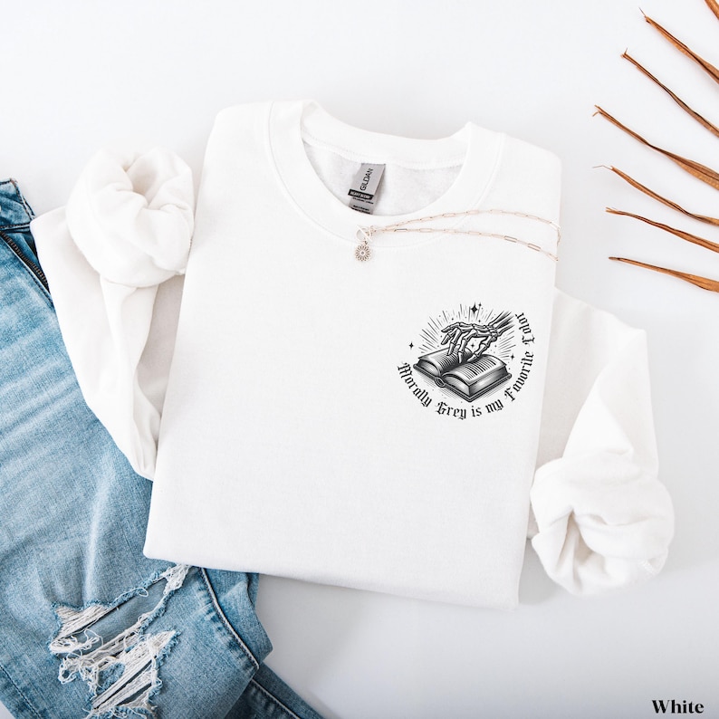May include: White sweatshirt with a crew neck and long sleeves. The graphic on the front features a skeleton hand reaching for an open book, with the text "Morally Grey is my Favorite Color" in a circular design. A silver necklace is draped across the sweatshirt.