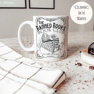 Op de afbeelding: Een witte keramische mok met een zwart-wit ontwerp. De mok heeft de tekst "The Banned Books Book Club" en een illustratie van een stapel boeken. De tekst "We stand with the banned" staat ook op de mok.