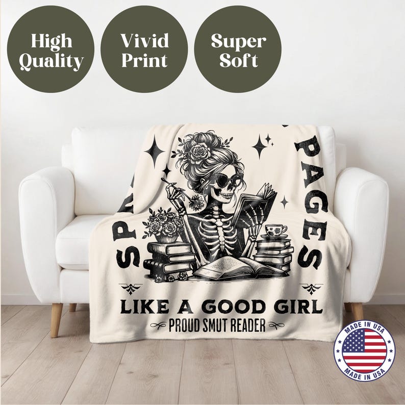 May include: A cream-colored throw blanket with a black and white illustration of a skeleton reading a book. The text on the blanket reads "SPA PAGES LIKE A GOOD GIRL PROUD SMUT READER".