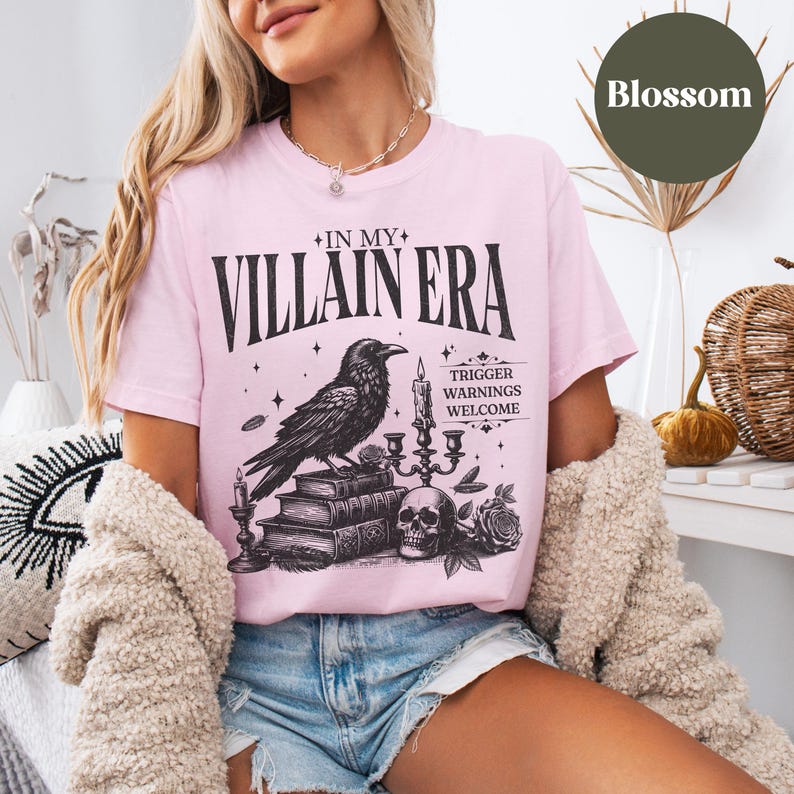 May include: Pink t-shirt featuring a black and white graphic print of a raven perched on a stack of books with a skull and candles. The text on the shirt reads "In My Villain Era" with the words "Trigger Warnings Welcome" below the graphic.