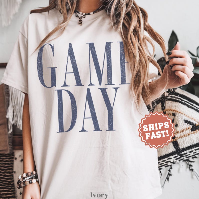 May include: An ivory short-sleeved t-shirt featuring the text GAME DAY in large, distressed navy blue capital letters. The shirt has a crew neckline and a relaxed fit. The word IVORY is printed in small letters at the bottom centre.