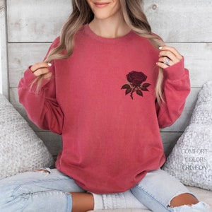 May include: A crimson red sweatshirt with a dark red rose graphic on the front. The sweatshirt is made of a soft, comfortable fabric and has a relaxed fit.