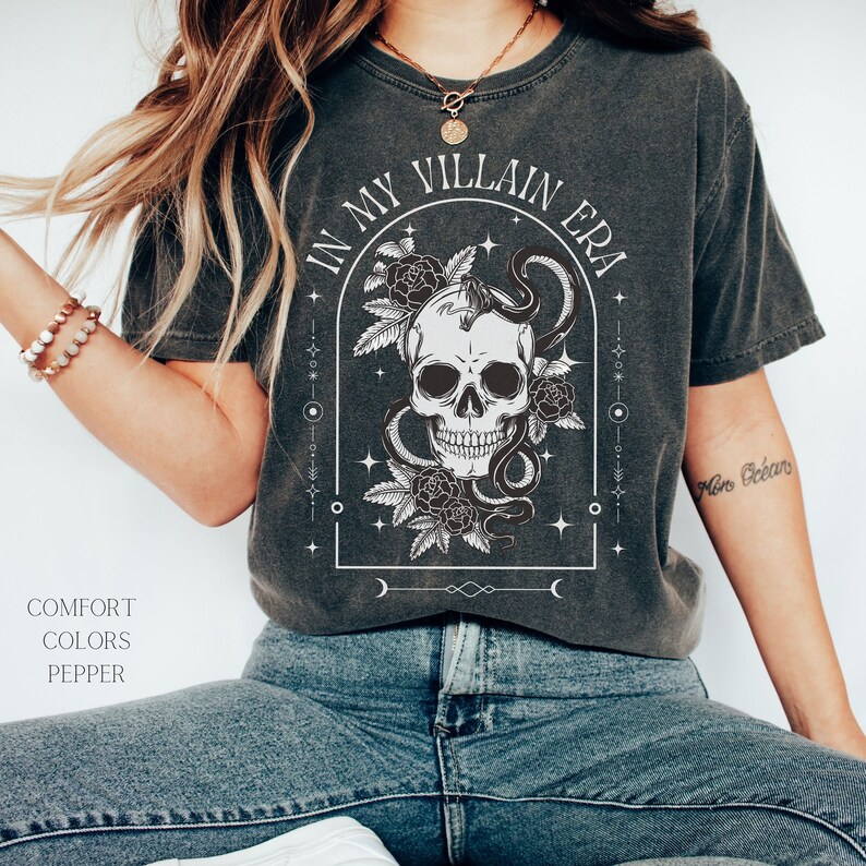 May include: A black t-shirt with a white graphic design of a skull with a snake wrapped around it. The graphic is surrounded by flowers and stars. The text "In My Villain Era" is written in a white arch above the skull.