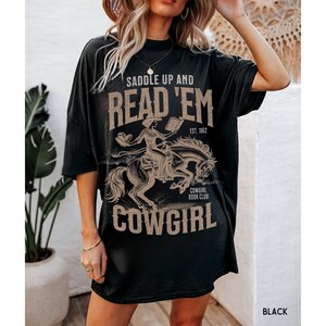 May include: Black oversized t-shirt with a graphic print of a cowgirl riding a horse and reading a book. The text on the shirt reads "Saddle Up and Read 'Em Cowgirl Book Club Est. 1862".