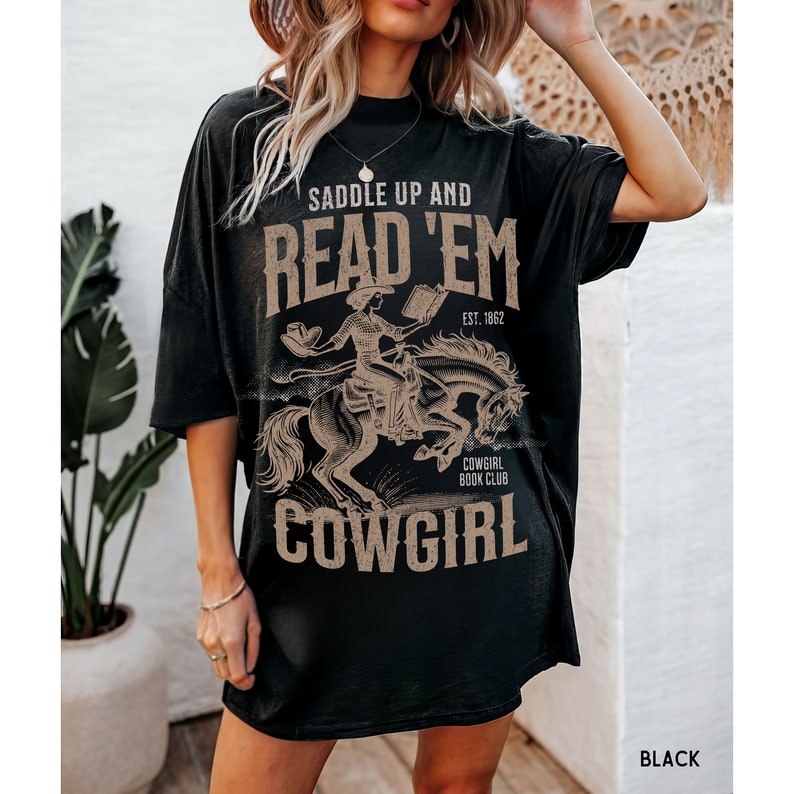 May include: Black oversized t-shirt with a graphic print of a cowgirl riding a horse and reading a book. The text on the shirt reads "Saddle Up and Read 'Em Cowgirl Book Club Est. 1862".