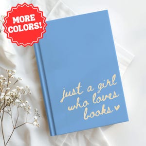 Può includere: Un diario blu chiaro con la scritta "just a girl who loves books" in oro. Un adesivo rosso nell'angolo in alto a sinistra dice "MORE COLORS!". Il diario è su una superficie bianca.