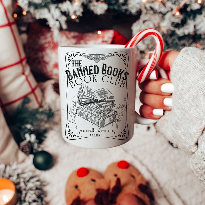 Op de afbeelding: Een witte keramische mok met een zwart-wit ontwerp met de tekst "The Banned Books Book Club" en een stapel boeken. De mok wordt vastgehouden door een hand met een snoepstok.