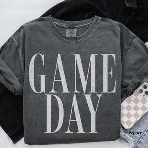May include: A charcoal grey short-sleeve t-shirt with a vintage-washed finish. The front features the text GAME DAY printed in large, distressed white capital letters. The shirt has a crew neckline and a standard fit, showcasing a casual aesthetic.