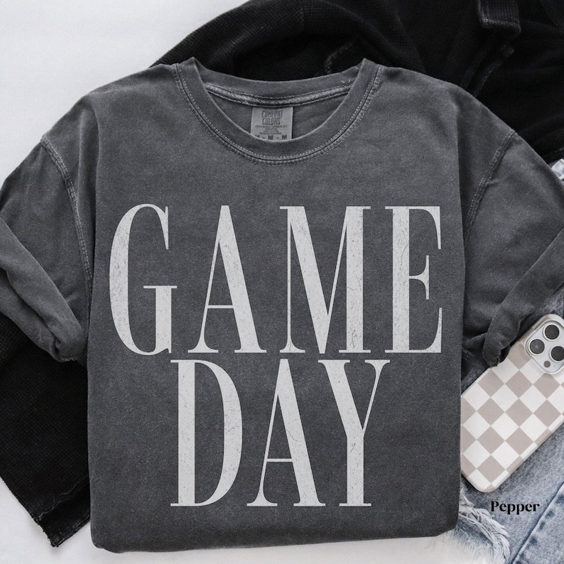 May include: A charcoal grey short-sleeve t-shirt with a vintage-washed finish. The front features the text GAME DAY printed in large, distressed white capital letters. The shirt has a crew neckline and a standard fit, showcasing a casual aesthetic.