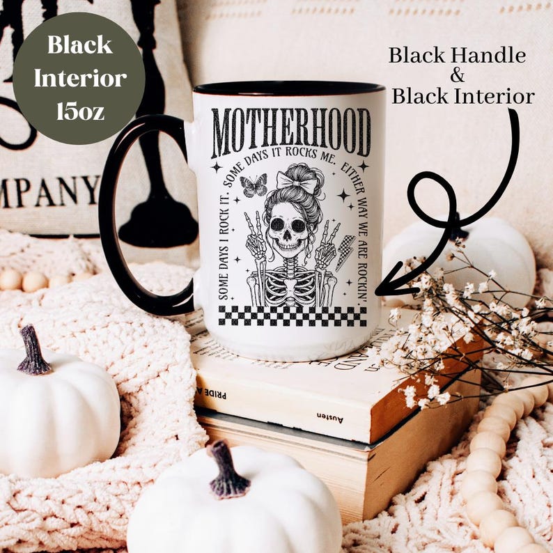 May include: White ceramic mug with a black handle and interior. The mug features a black and white illustration of a skeleton with a skull face wearing a bow and holding up peace signs. The text "Motherhood Some Days I Rock It. Some Days It Rocks Me. Either Way We Are Rockin'" is printed on the mug.