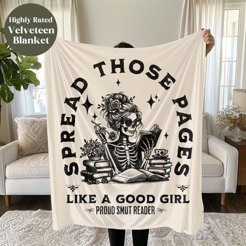 May include: A white throw blanket with a black and white illustration of a skeleton reading a book. The text on the blanket reads "Spread Those Pages Like A Good Girl Proud Smut Reader".
