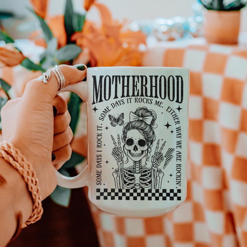 May include: White ceramic mug with a black and white graphic of a skeleton with a crown and the text "Motherhood. Some days I rock it. Some days it rocks me. Either way we are rockin'."