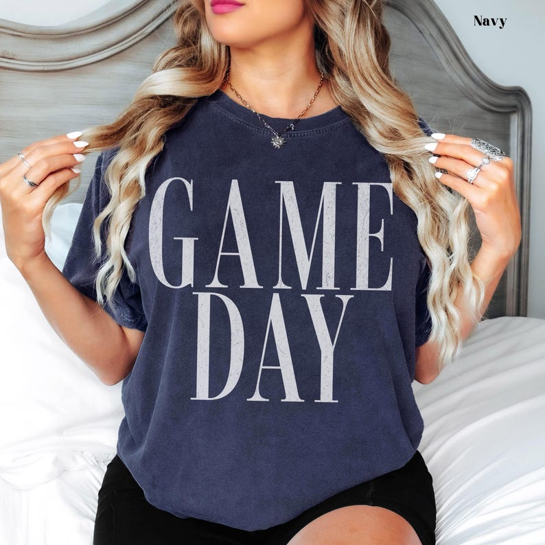 May include: A navy blue short-sleeved t-shirt featuring a distressed, vintage-style white graphic print on the front which reads "GAME DAY" in large, bold, capital letters. The garment has a crew neckline and a relaxed, comfortable fit.