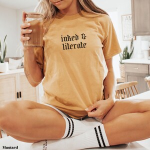 May include: Mustard-colored t-shirt with the text "inked & literate" in black gothic font. The shirt is paired with white socks featuring black stripes. A clear glass with a brown beverage is held in the hand.