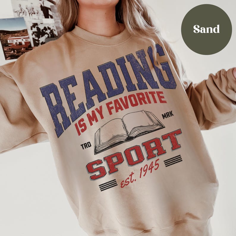 May include: A tan sweatshirt with the text "Reading is my favorite sport est. 1945" in red, blue, and black lettering. The text is surrounded by a graphic of an open book.