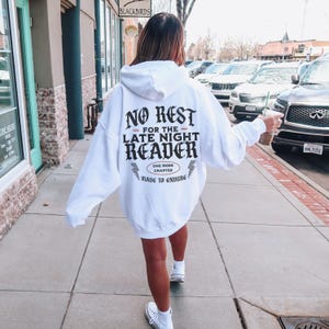 Peut inclure: Un sweat &agrave; capuche blanc avec l'inscription "NO REST FOR THE LATE NIGHT READER" en lettres gothiques noires. Le sweat &agrave; capuche comporte &eacute;galement le texte "ONE MORE CHAPTER MADE TO ENDURE" et des &eacute;clairs. La personne marche sur un trottoir.