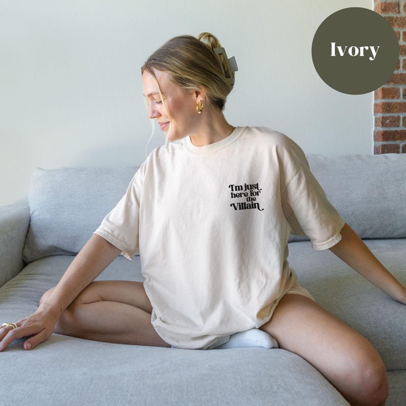 Puede incluir: Una mujer lleva una camiseta beige claro con el texto "I'm just here for the Villain" en negro. La camiseta es de corte holgado y tiene cuello redondo.