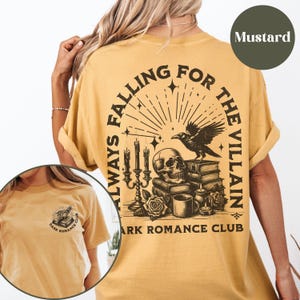 May include: A mustard yellow t-shirt with a black graphic print of a skull, raven, and books. The text "Always Falling For The Villain" is printed in a circle around the graphic. The text "Dark Romance Club" is printed below the graphic.
