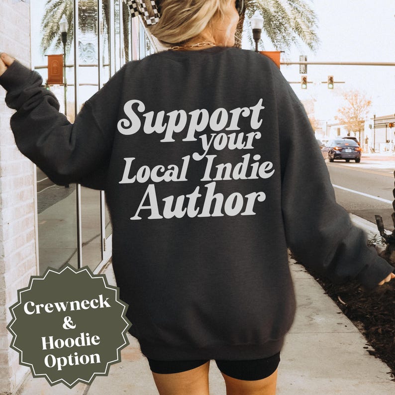 May include: Dark gray crewneck sweatshirt with the text "Support your Local Indie Author" in white script across the back. A small graphic at the bottom left corner reads "Crewneck & Hoodie Option".