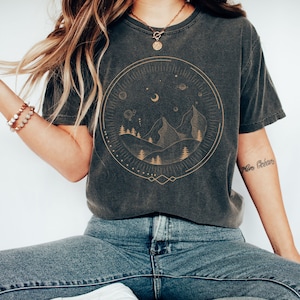 Puede incluir: Una camiseta gris oscura con un gr&aacute;fico dorado de una cordillera, luna y estrellas dentro de un c&iacute;rculo. El gr&aacute;fico tiene un estilo vintage y celestial.