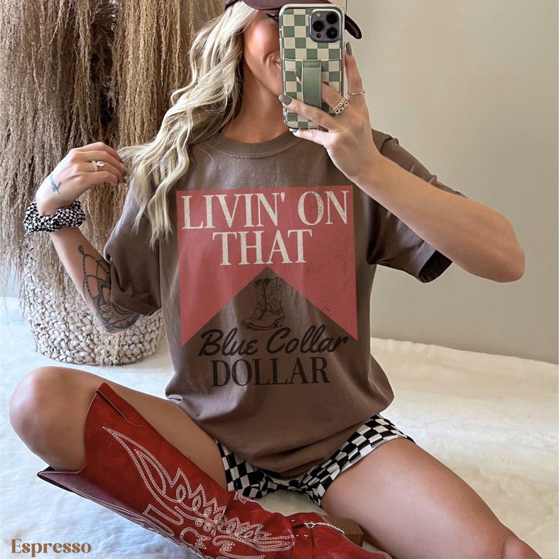 May include: A brown t-shirt with a red and white graphic that says "Livin' on That Blue Collar Dollar". The graphic features a vintage-style illustration of a work boot. The t-shirt is being worn by a person with long blonde hair and a brown hat.