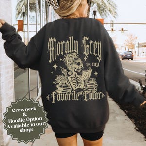 May include: Dark gray sweatshirt with the text "Morally Grey is my Favorite Color" in a gothic font. The design features a skeleton reading a book, with flowers and stars. The sweatshirt also has a "Crewneck & Hoodie Option Available in our shop!" text.