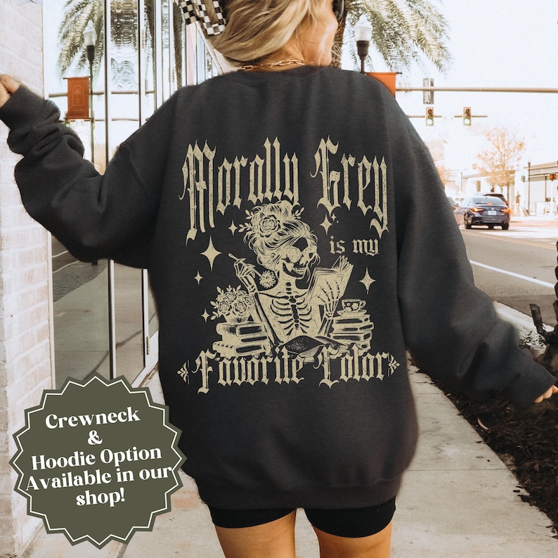 May include: Dark gray sweatshirt with the text "Morally Grey is my Favorite Color" in a gothic font. The design features a skeleton reading a book, with flowers and stars. The sweatshirt also has a "Crewneck & Hoodie Option Available in our shop!" text.