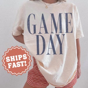 May include: A cream-coloured short-sleeve t-shirt featuring the text "GAME DAY" printed in large, distressed navy blue capital letters across the chest. The shirt is paired with red and white gingham patterned shorts.