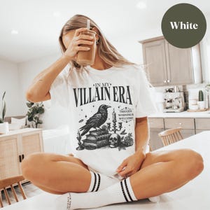 May include: White t-shirt featuring a black and white graphic of a raven, candles, and books. The text on the shirt reads "In My Villain Era" and "Trigger Warnings Welcome".