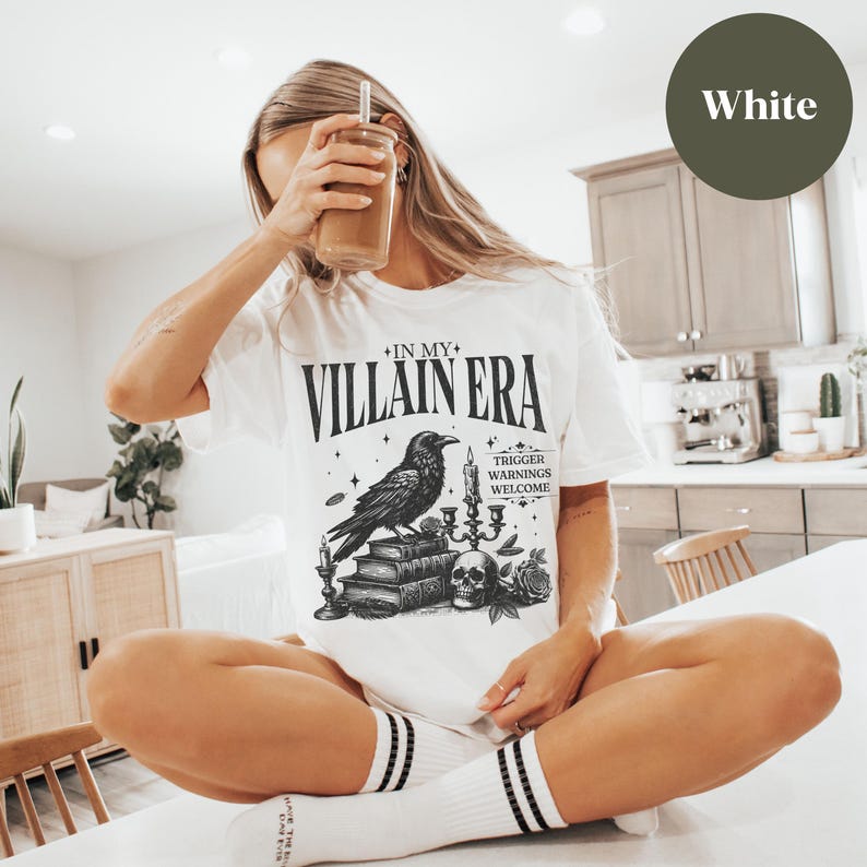 May include: White t-shirt featuring a black and white graphic of a raven, candles, and books. The text on the shirt reads "In My Villain Era" and "Trigger Warnings Welcome".