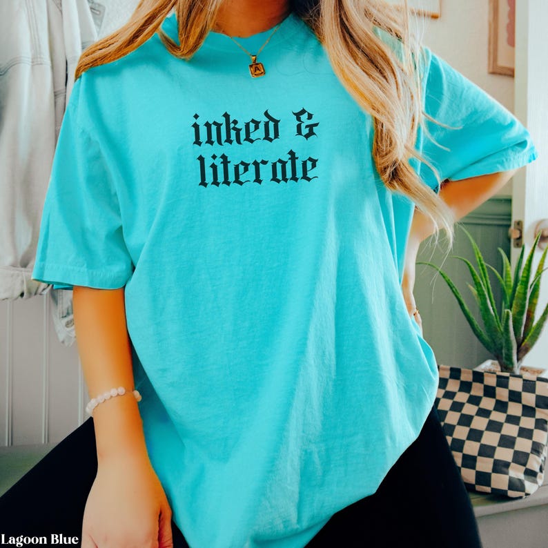 May include: Lagoon blue t-shirt with the words "inked & literate" in black gothic font. The short-sleeved shirt is made of a soft material and is suitable for casual wear.