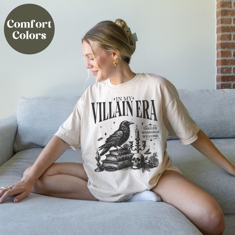 May include: A beige t-shirt with a black graphic print of a raven, candles, books, a skull, and roses. The text on the shirt reads "In My Villain Era" and "Trigger Warnings Welcome".
