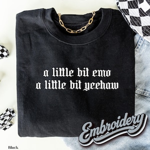 May include: Black sweatshirt with the text "a little bit emo a little bit yeehaw" in white gothic font. A gold chain necklace is draped around the neckline. The word "Embroidery" is printed on the bottom right.