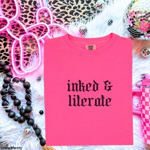 May include: A bright pink t-shirt with the words "inked & literate" in black gothic font. The shirt is folded neatly on a white fluffy surface, with disco balls and leopard print accents in the background.