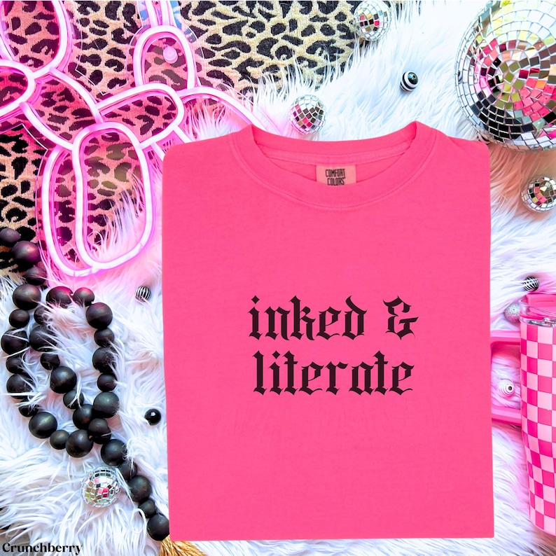 May include: A bright pink t-shirt with the words "inked & literate" in black gothic font. The shirt is folded neatly on a white fluffy surface, with disco balls and leopard print accents in the background.