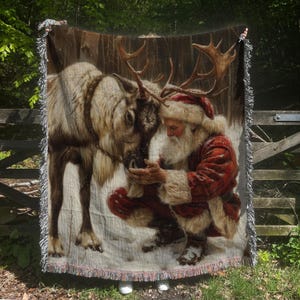 May include: A woven throw blanket featuring a detailed illustration of Santa Claus with a white beard and red coat kneeling in the snow to touch the nose of a large reindeer with antlers. The blanket has a fringed edge on the top and bottom.