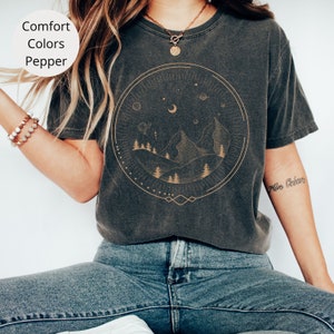 Puede incluir: Una camiseta gris oscura con un gr&aacute;fico de una cordillera, luna y estrellas dentro de un c&iacute;rculo. El gr&aacute;fico es de color dorado.
