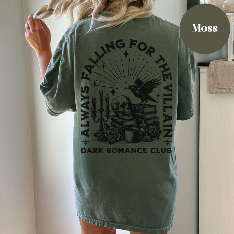 May include: A green t-shirt with a black graphic print that says "Always Falling For The Villain" and "Dark Romance Club". The graphic features a skull, a raven, candles, and books.
