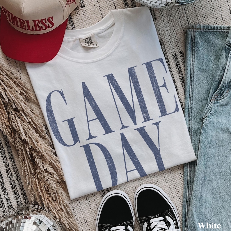 May include: A white short-sleeved t-shirt featuring the text GAME DAY in a distressed blue font. The garment is shown with a beige and red baseball cap bearing the word TIMELESS, a pair of blue denim jeans, and a pair of black trainers.