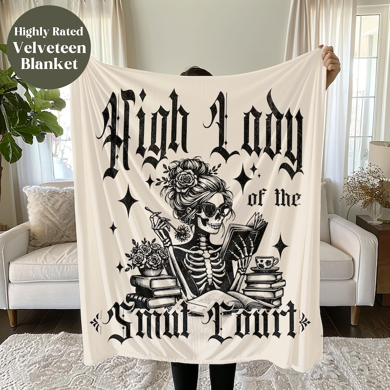 May include: A white throw blanket with a black and white illustration of a skeleton woman reading a book. The text "High Lady of the Snow Court" is printed on the blanket.