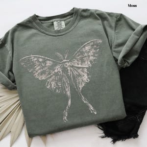 Puede incluir: Una camiseta de manga corta color verde musgo con un gran estampado gr&aacute;fico blanco desgastado de una polilla con alas largas y extendidas. La prenda cuenta con un cuello redondo y una etiqueta de marca visible en el interior del cuello.