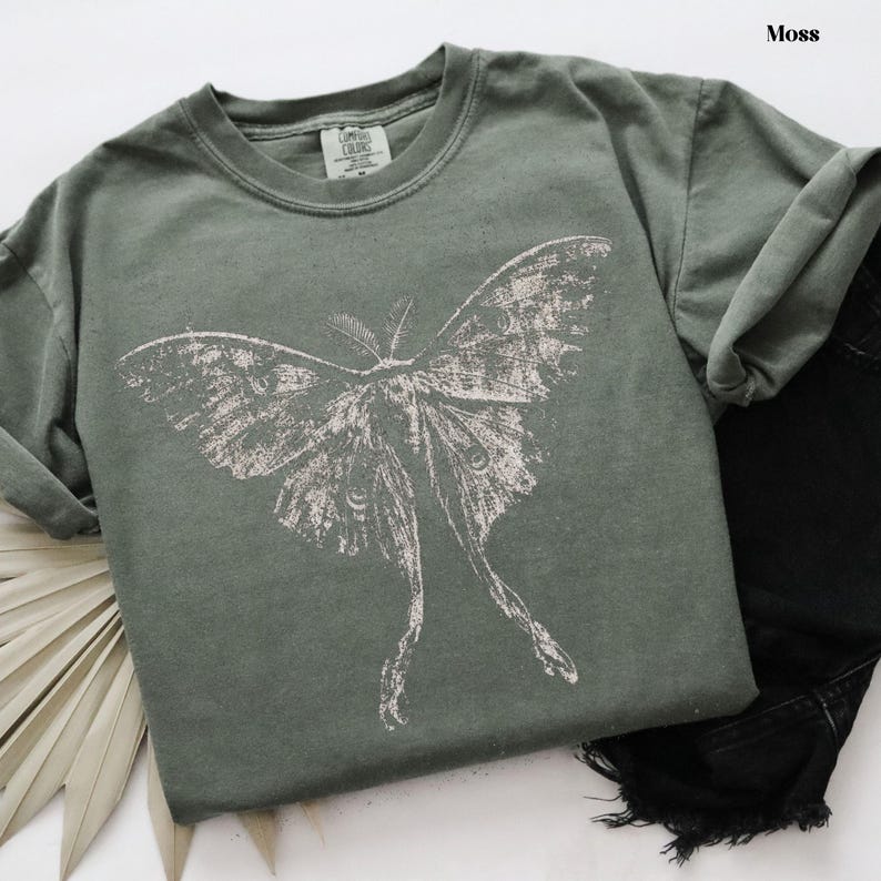 Puede incluir: Una camiseta de manga corta color verde musgo con un gran estampado gr&aacute;fico blanco desgastado de una polilla con alas largas y extendidas. La prenda cuenta con un cuello redondo y una etiqueta de marca visible en el interior del cuello.