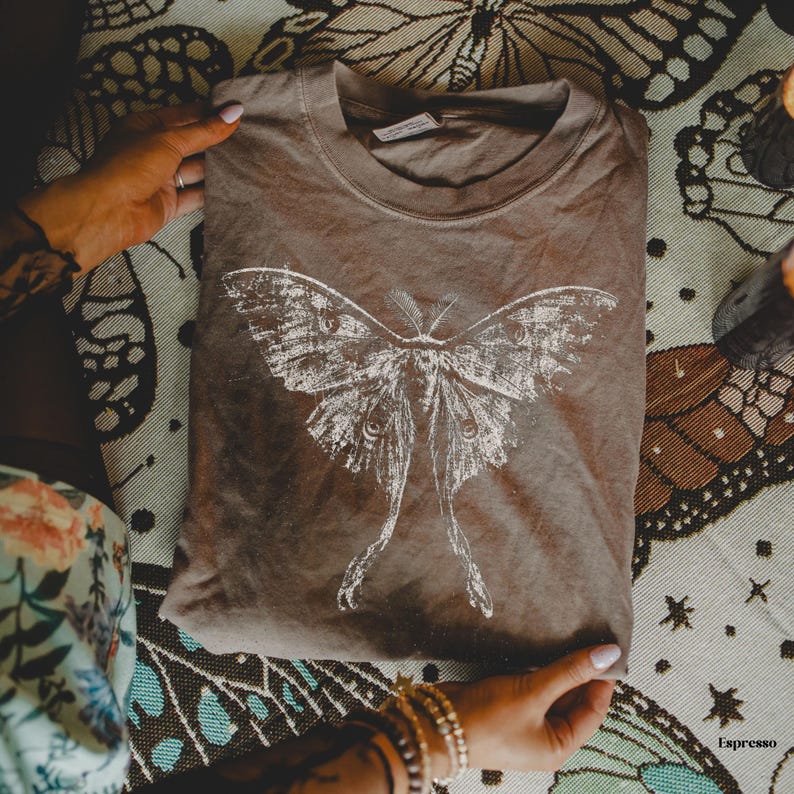 Puede incluir: Una camiseta marr&oacute;n con un gran estampado gr&aacute;fico blanco de estilo desgastado que representa una polilla con alas largas y alargadas. La camiseta tiene cuello redondo y una etiqueta visible en el interior del cuello.