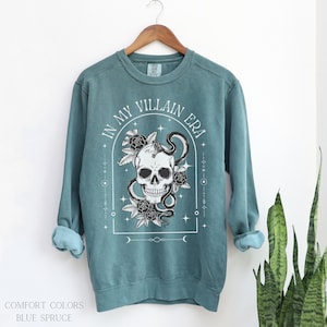 May include: A teal blue sweatshirt with a white graphic print of a skull with a snake wrapped around it. The graphic also includes roses and the text "In My Villain Era".