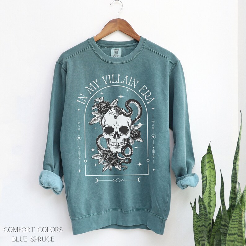 May include: A teal blue sweatshirt with a white graphic print of a skull with a snake wrapped around it. The graphic also includes roses and the text "In My Villain Era".