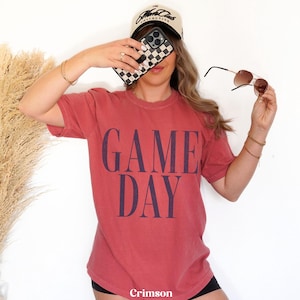 May include: A short-sleeved, crew-neck t-shirt in a faded crimson red colour. The shirt features the words GAME DAY printed in large, bold, dark navy blue capital letters across the centre of the chest. The fabric has a soft, worn-in appearance.