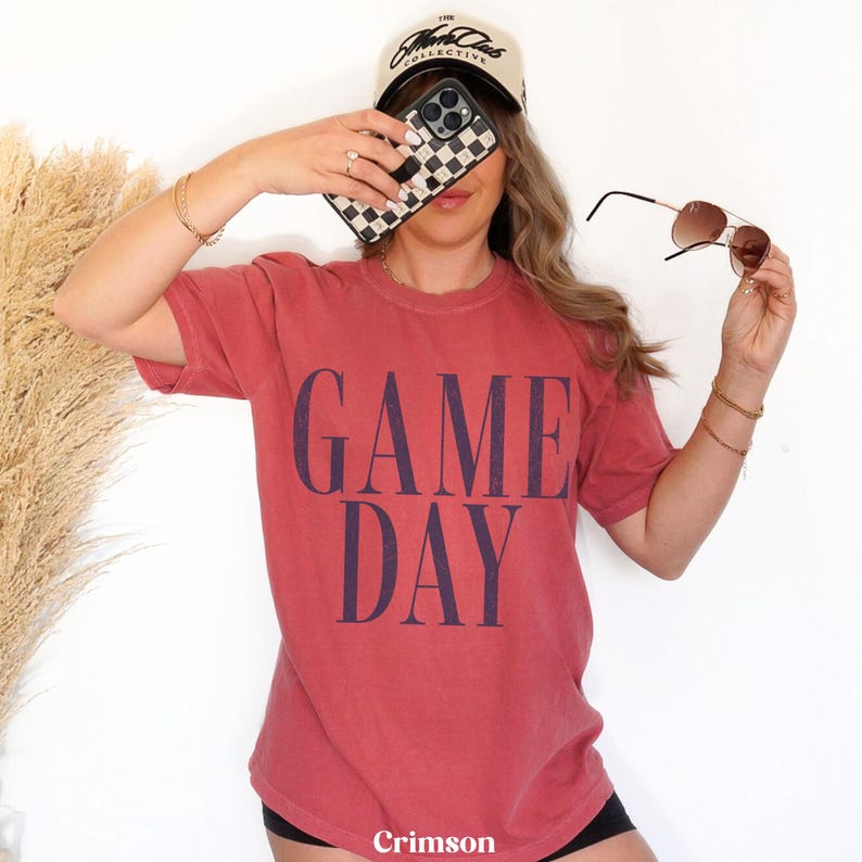 May include: A short-sleeved, crew-neck t-shirt in a faded crimson red colour. The shirt features the words GAME DAY printed in large, bold, dark navy blue capital letters across the centre of the chest. The fabric has a soft, worn-in appearance.