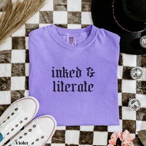 May include: A lavender t-shirt with the words "inked & literate" in black gothic font. The shirt is paired with white sneakers and a black hat. The background has a checkered pattern.