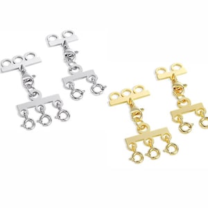 May include: Two sets of jewellery clasps in silver and gold. Each clasp has three small rings and a lobster claw clasp. The clasps are designed to connect multiple strands of necklaces or bracelets. The background is white.