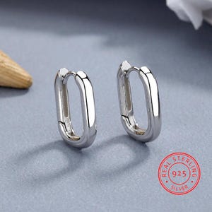 May include: A pair of polished silver hoop earrings with an elongated oval shape and a hinged closure mechanism. A circular red stamp in the corner features the text Real Sterling 925 Silver, indicating the metal composition.